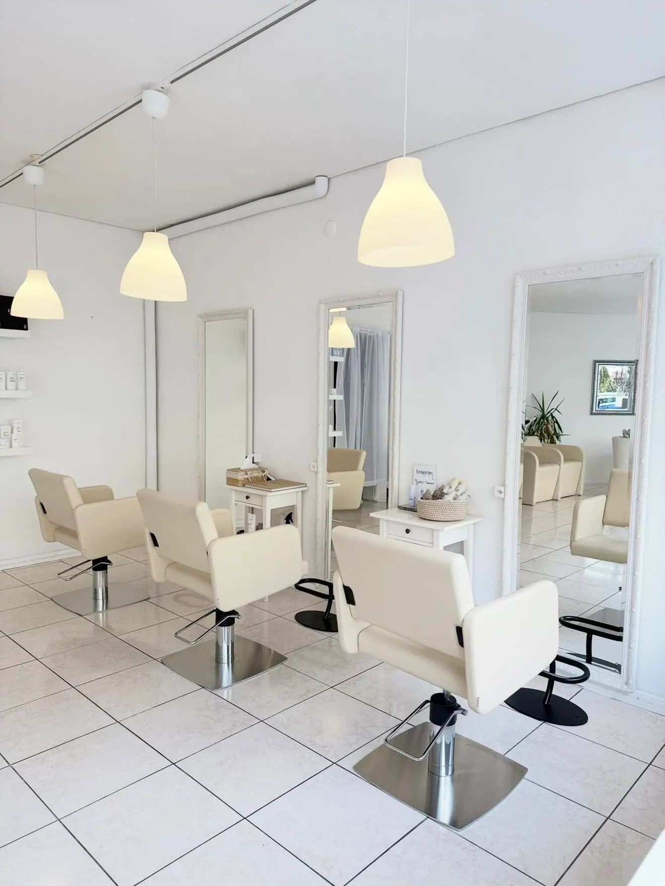 Der Salon von innen: drei cremefarbene Stuehle vor weiss gerahmten Spiegeln, warmes Licht aus Pendelleuchten.